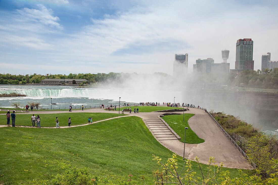 'Visitors Stand Behind A Railing Overlooking Horseshoe Falls At Niagara Falls State Park, In New York State With The Town Of Niagara Falls, Ontario In The Background; Niagara Falls, New York, United States Of America'