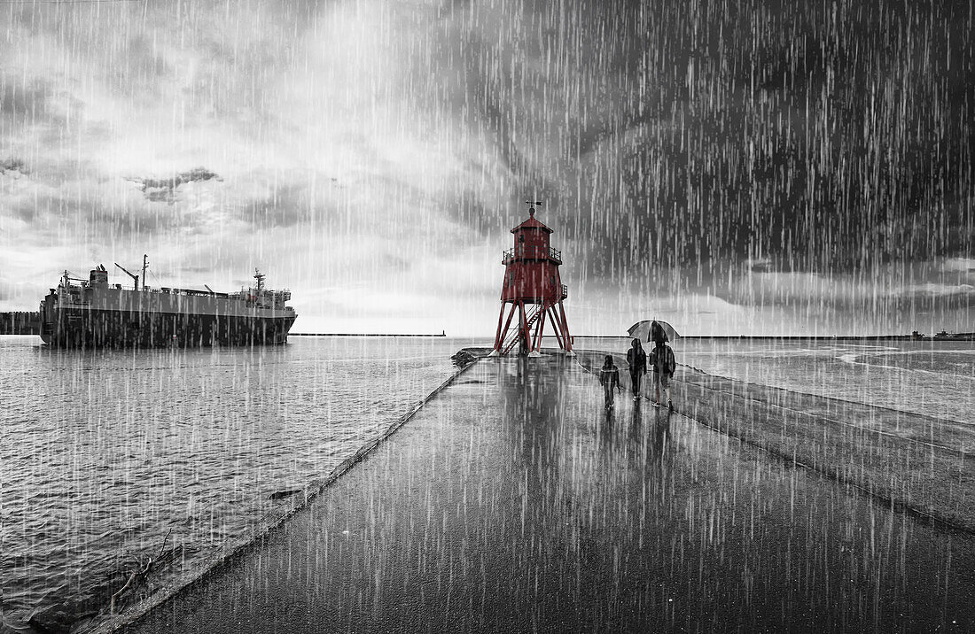 'A family walking in the pouring rain along harbour beach with Groyne Lighthouse and a ship; South Shields, Tyne and Wear, England'