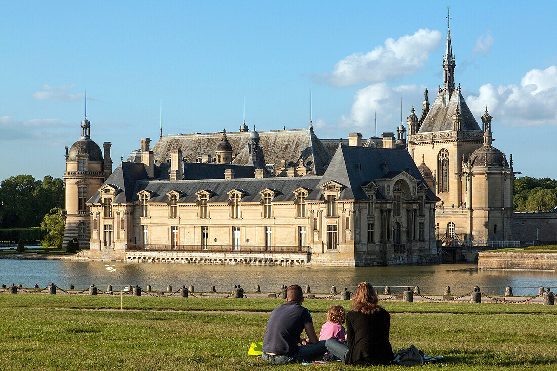 Family sitting on the lawn across from the chateau de chantilly and its moat, oise (60), france