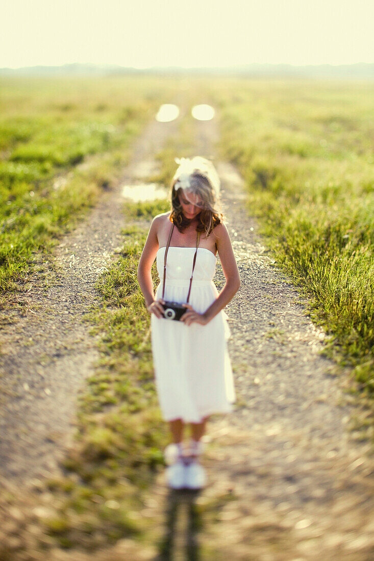 A Woman In A White Dress Standing On A Dirt Road With A Camera Around Her Neck