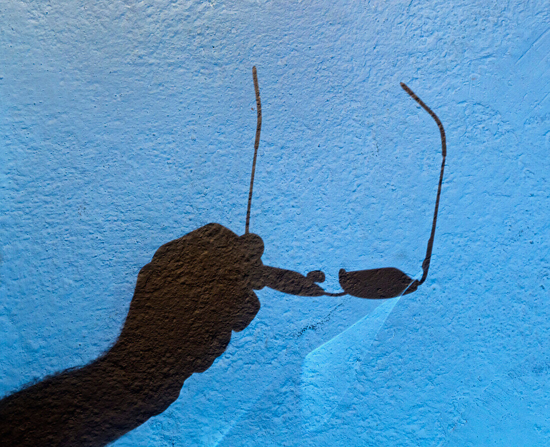'Shadow Of Hand Holding Eyeglasses Against A Blue Wall; Tarifa, Spain'