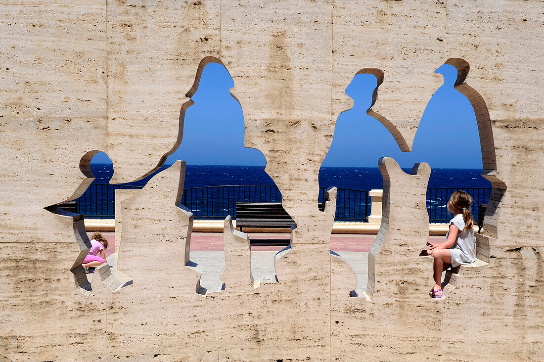 A young girl sitting on sculpture in front of Mediterranean sea in Valletta, Malta, Europe