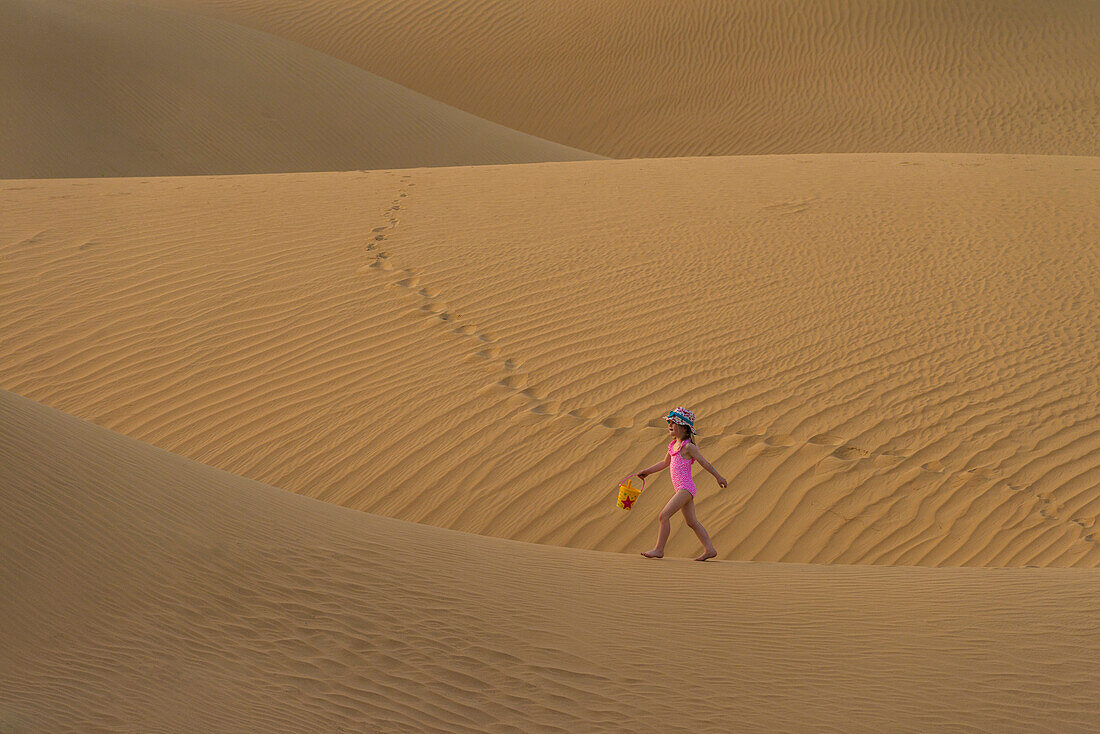 'Girl in pink swimsuit with bucket and spade walking across sand dunes at dusk; Dubai, United Arab Emirates'