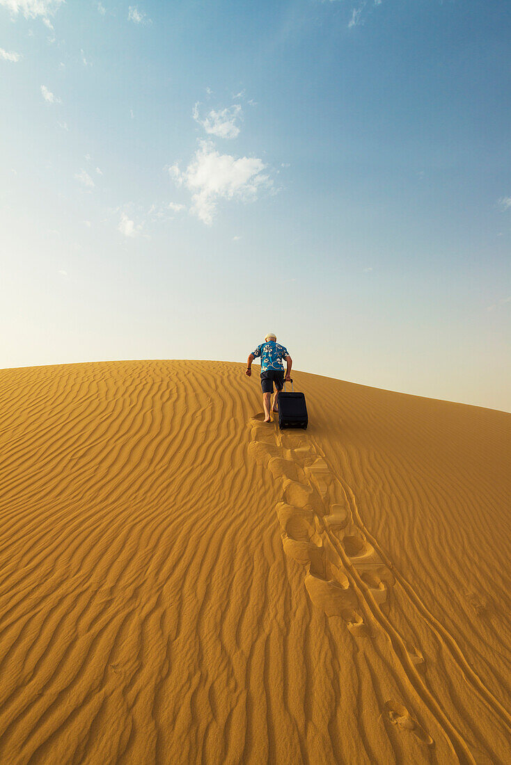 'Barefoot man with suitcase walking up sand dune; Dubai, United Arab Emirates'