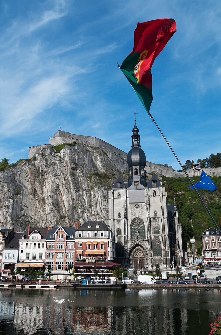 'A church and other buildings along the water and flags flying; Dinant, Belgium'