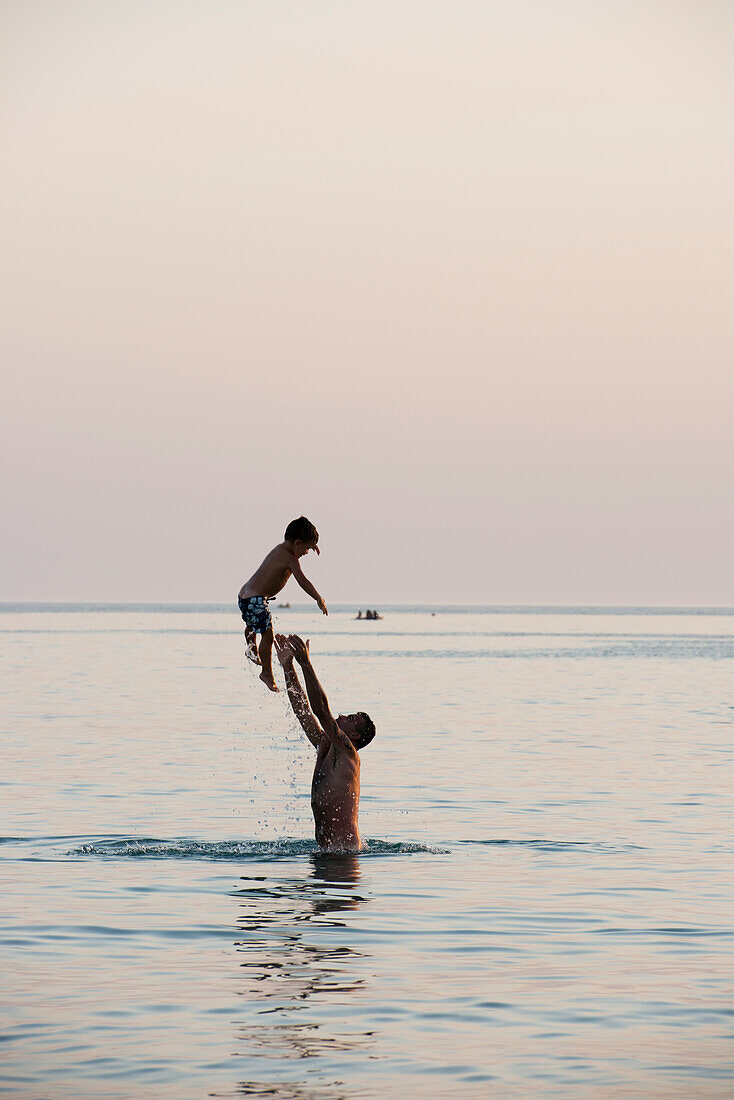 'A father standing in the water and throwing his son into the air; Puglia, Italy'