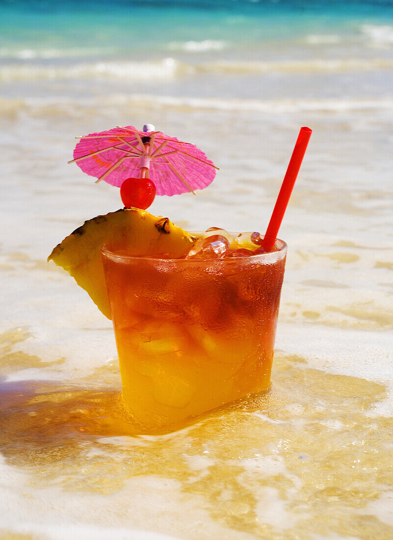 A Mai Tai Garnished With Pinapple And A Cherry, Sitting In Shallow Water On The Beach.
