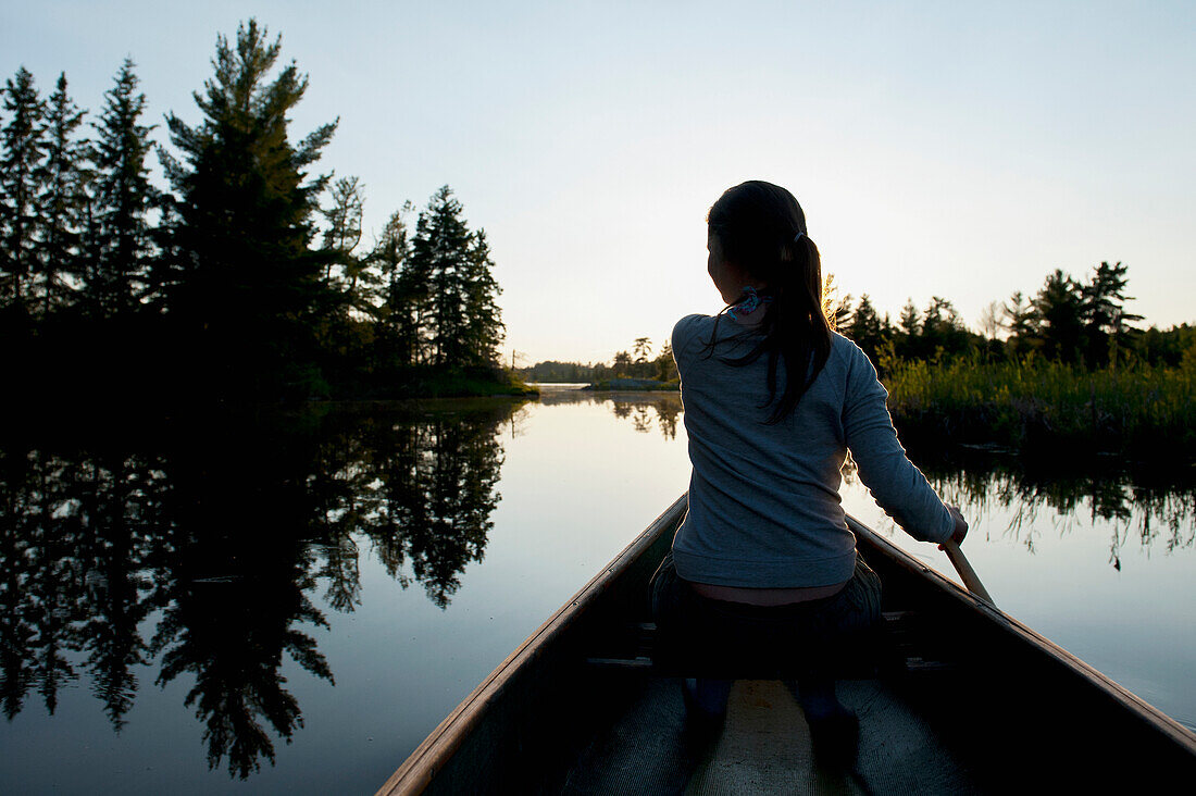 'A Girl Paddles A Boat On A Tranquil Lake At Sunset; Lake Of The Woods, Ontario, Canada'