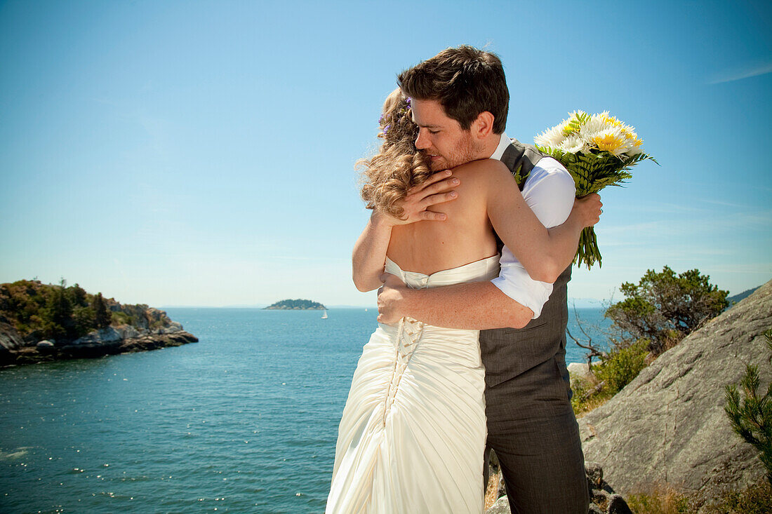 'A Bride And Groom Embrace At The Water's Edge In Whytecliff Park; Vancouver, British Columbia, Canada'
