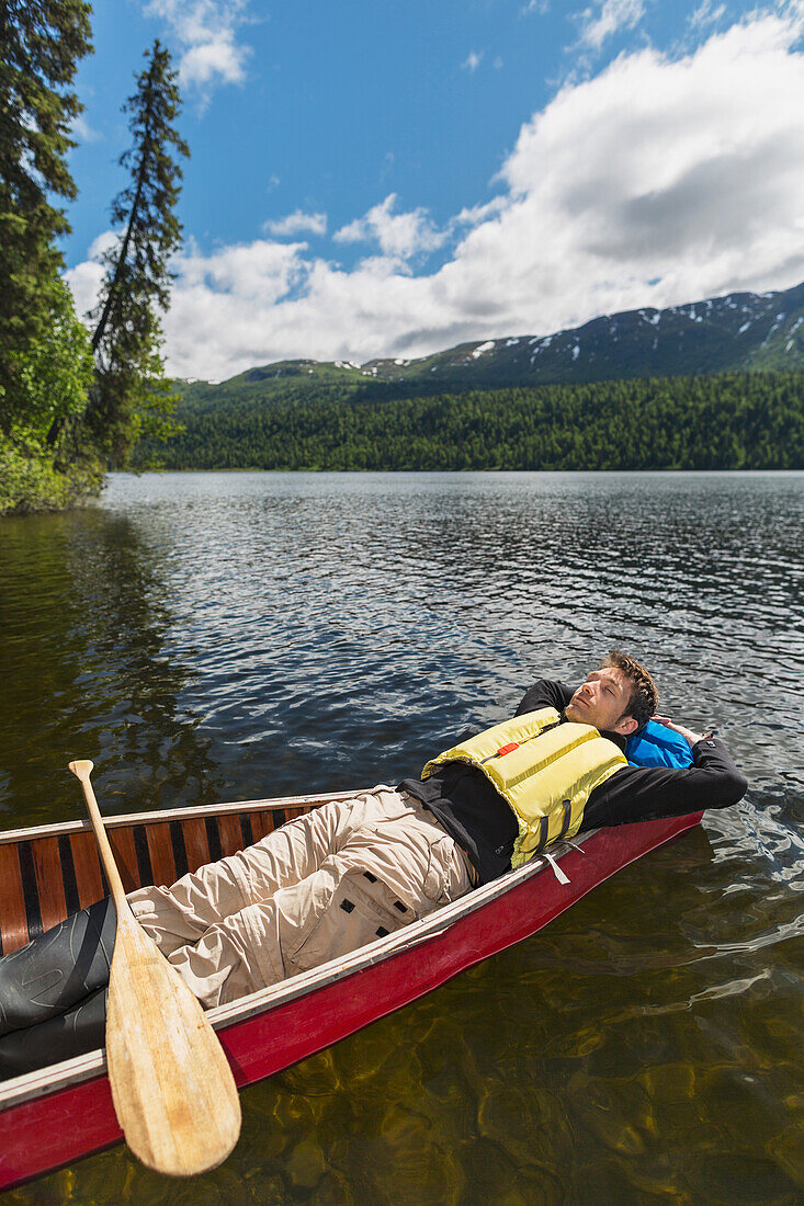 'Man reclined in canoe and resting in the sunshine on Byers Lake with forested foothills in the background, Byers lake campground, Denali State Park; Alaska, United States of America'