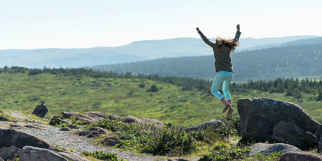 'A young woman jumps from a rock with arms in the air; St. John's, Newfoundland and Labrador, Canada'