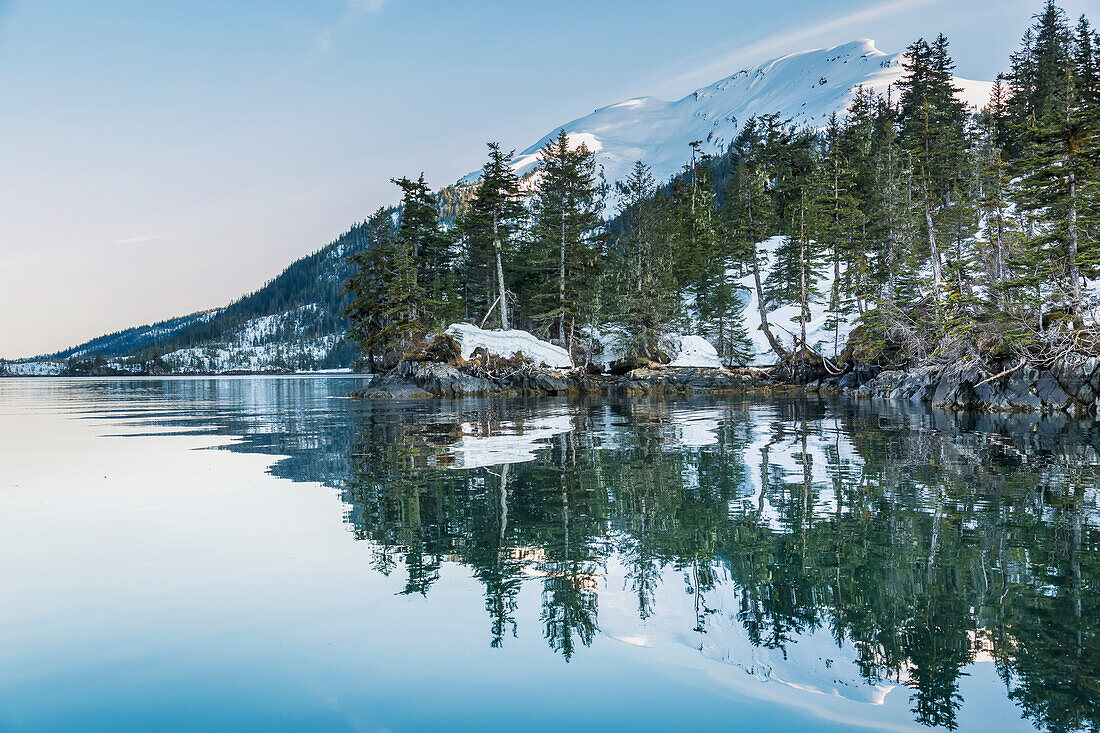 'Snow covered mountains and shoreline with trees reflected in the waters of Harrison Lagoon, Chugach National Forest; Alaska, United States of America'