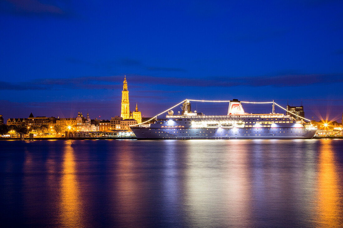 Cruise ship MS Deutschland (Reederei Peter Deilmann) moored along the Scheldt river with Cathedral of Our Lady church tower and skyline at night, Antwerp, Flemish Region, Belgium