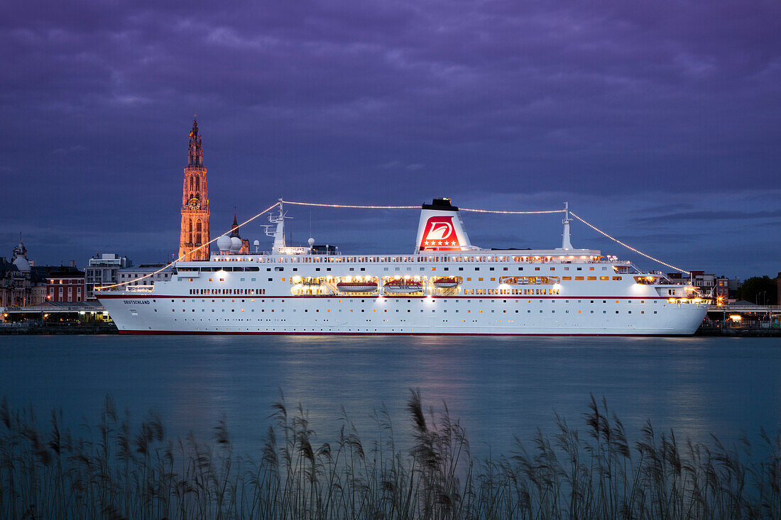 Cruise ship MS Deutschland (Reederei Peter Deilmann) moored along the Scheldt river with Cathedral of Our Lady church tower at dusk, Antwerp, Flemish Region, Belgium