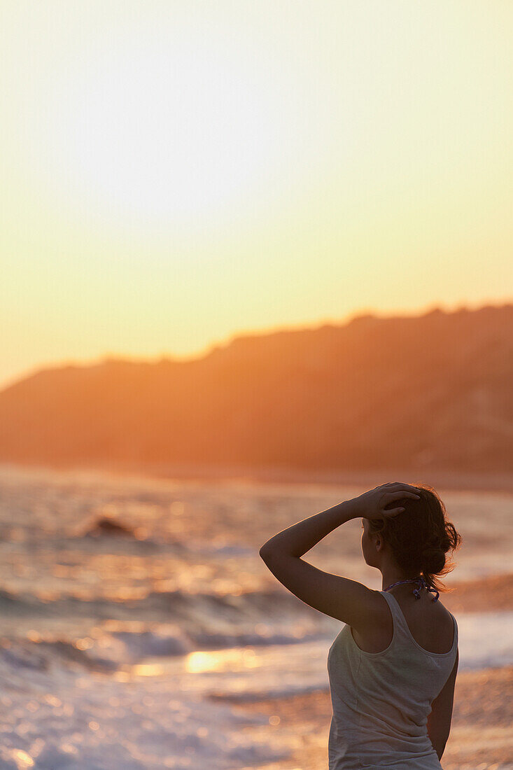 'A young woman stands at the water's edge watching the sunset;Aphrodite bay, cyprus'