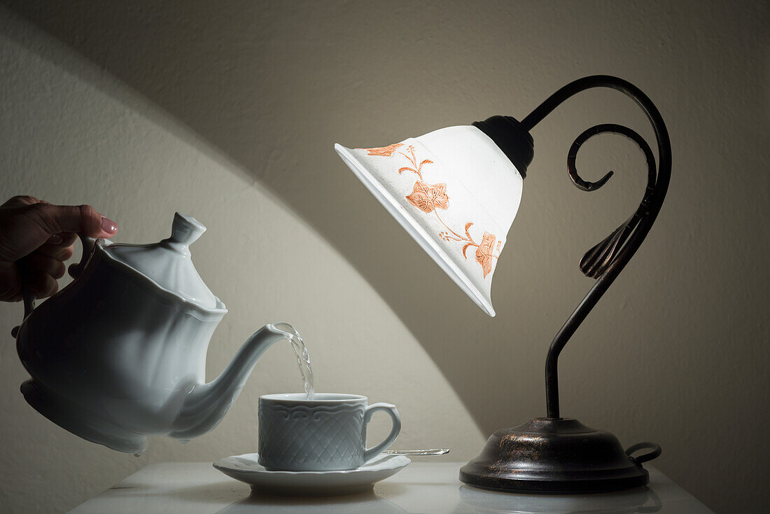 'A Lamp Shining On A Hand Holding A Teapot Pouring Tea Into A Teacup; Sestri Levante, Emilia-Romagna, Italy'
