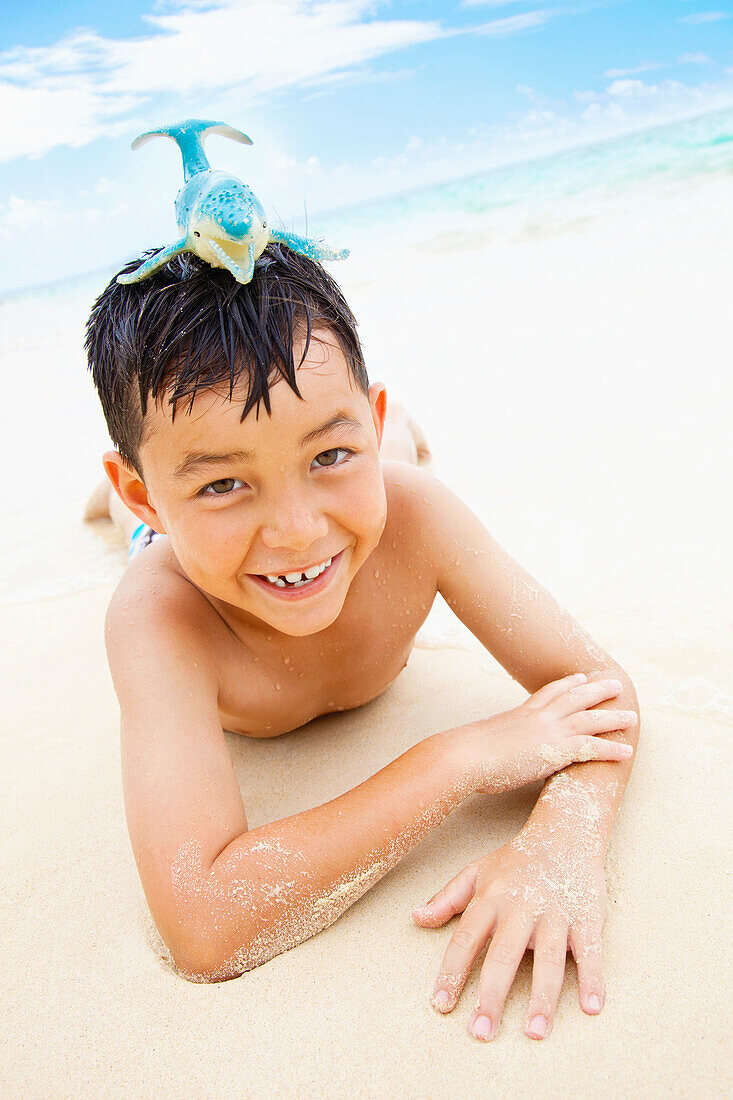 'A Boy Laying On The Sand At The Water's Edge With A Toy Whale On His Head; Kailua, Oahu, Hawaii, United States Of America'