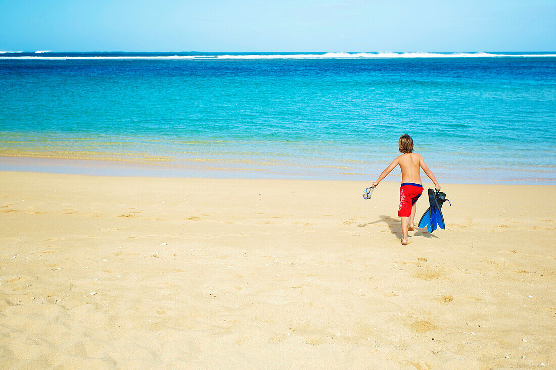 'A boy running out to the ocean across the beach with snorkelling gear; Kauai, Hawaii, United States of America'