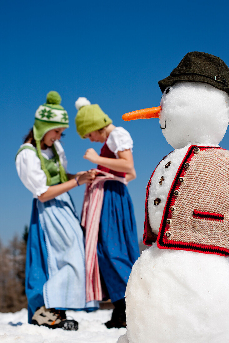 Two girls wearing dirndls, snowman in foreground, Frauenalpe, Murau, Styria, Austria