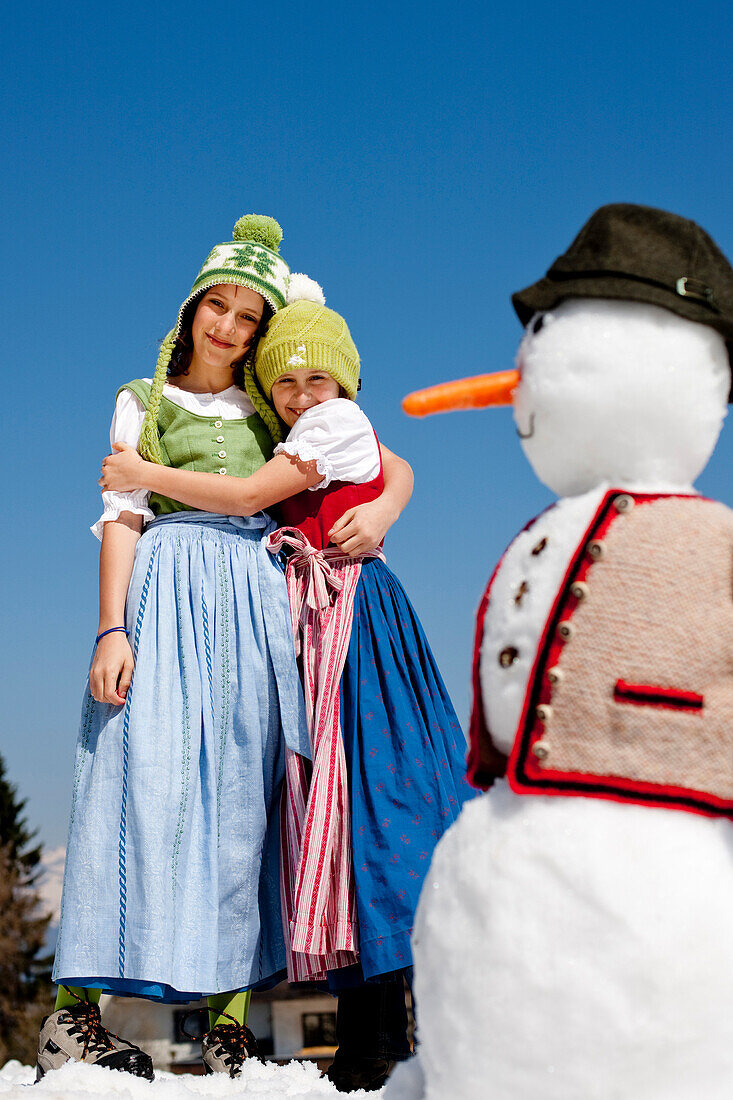 Zwei Mädchen im Dirndl umarmen sich, Schneemann im Vordergrund, Frauenalpe, Murau, Steiermark, Österreich