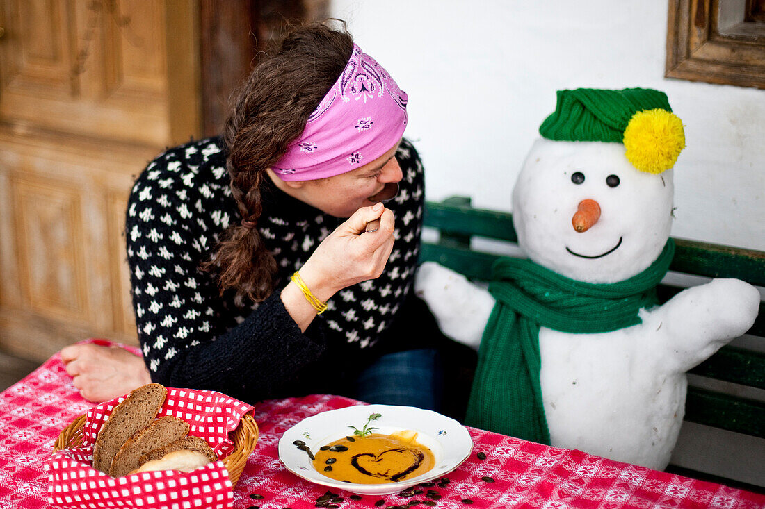 Woman sitting on a bench beside a snowman while eating pumpkin cream soup, Styria, Austria