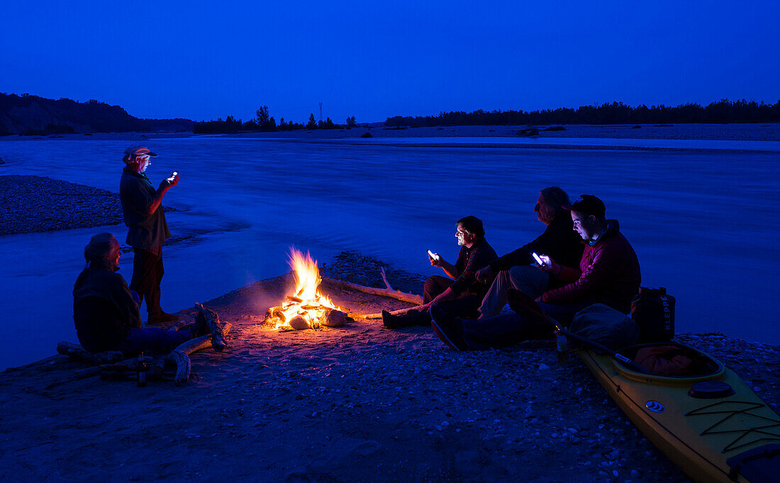 Group of paddlers with smart phones at camp fire, Tagliamento river, Friuli, Italy