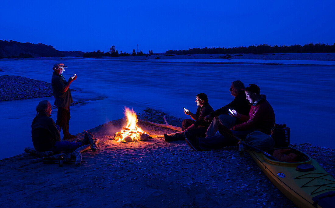 Gruppe Paddler mit Smartphones am Lagerfeuer am Ufer des Tagliamento, Friaul, Italien