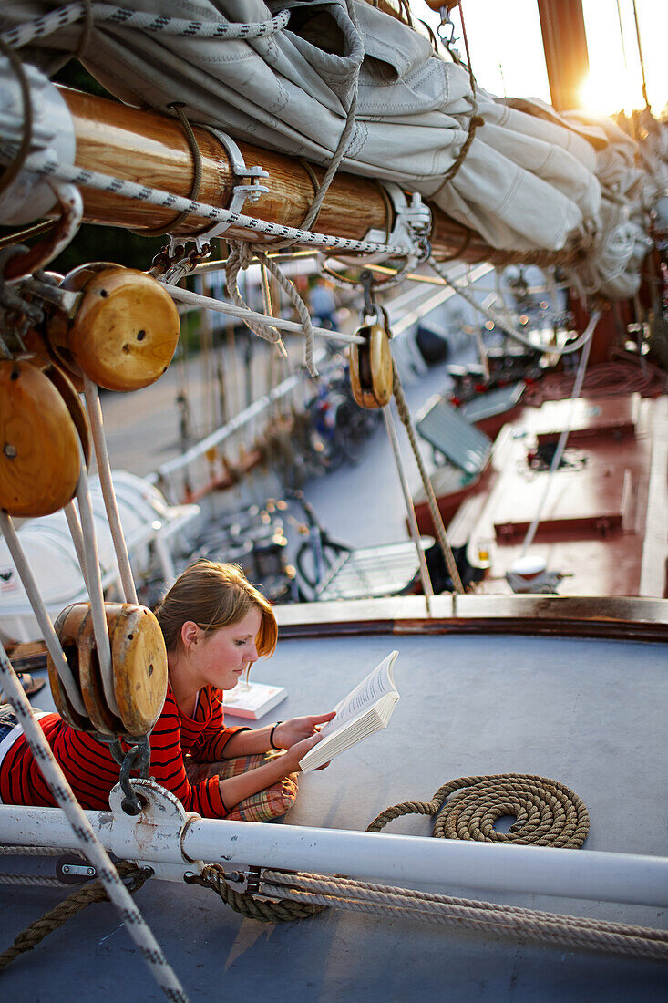 Teenager lying on deck of sailing ship while reading a book, Lauterbach, Putbus, insland of Ruegen, Mecklenburg-Western Pomerania, Germany
