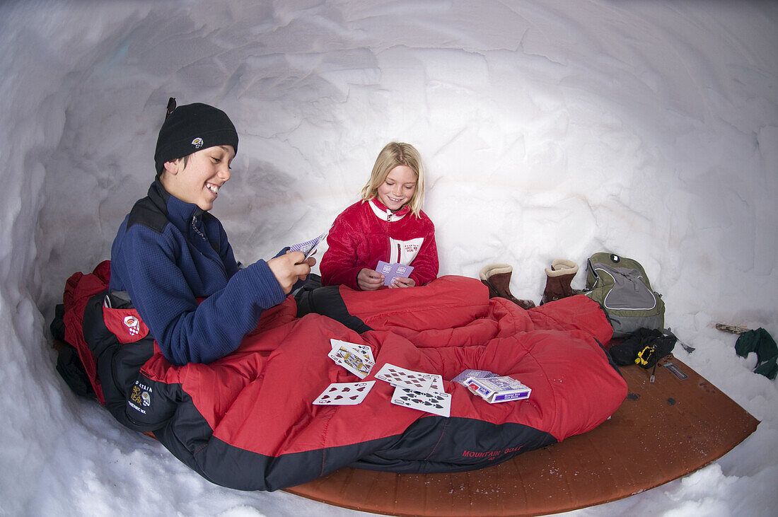 Boy and girl playing cards in sleeping bags whild camping in snowcave on Red Mountain Pass, San Juan National Forest, Colorado.