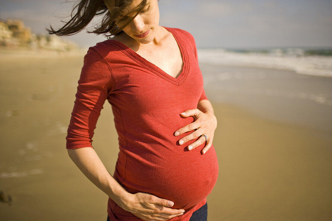 A pregnant woman hugs her belly contently, while facing into the wind on a sunny day at the beach in California.