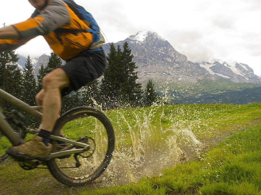 A mountain biker is driving through a mud pool on the bike trail towards First. In the background the Eiger mountain.