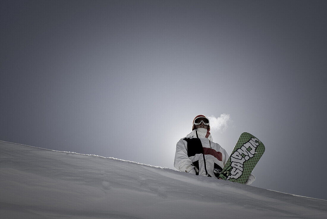 Portrait of backlit snowboarder standing with board on horizon.