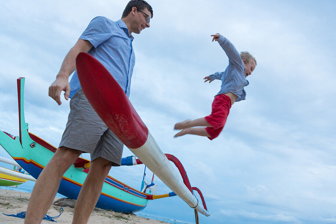 Vater und Sohn spielen am Strand, Junge springt vom Boot, traditionelles Fischerboot, fliegen, Kleinkind, 3 Jahre alter Junge, Palme, Elternzeit in Asien, Europäer, Deutsche, Westler, Familie, MR, Sanur, Bali, Indonesien