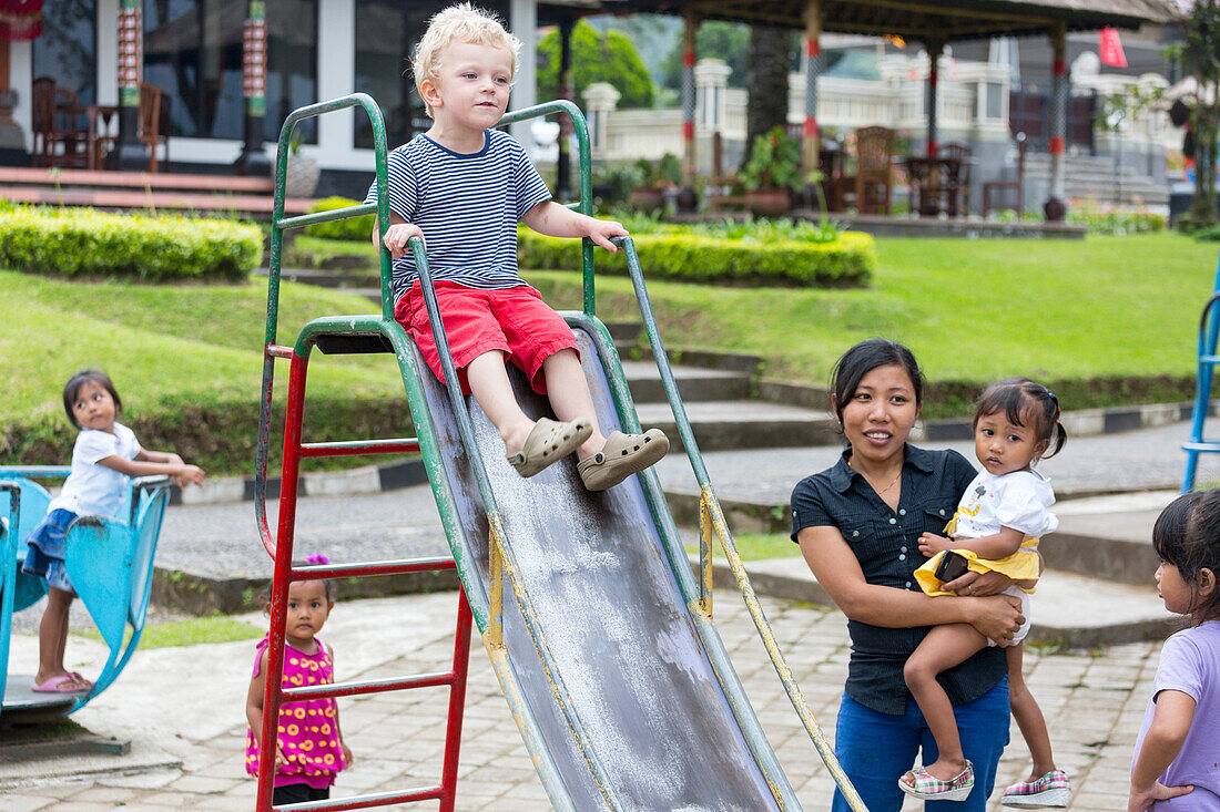 German boy on a slide, playing with Balinese kids and women, playground at the temple Pura Ulun Danau Bratan, intercultural contact, meeting locals, family travel in Asia, parental leave, German, European, MR, Ubud, Bedugul, Bali, Indonesia