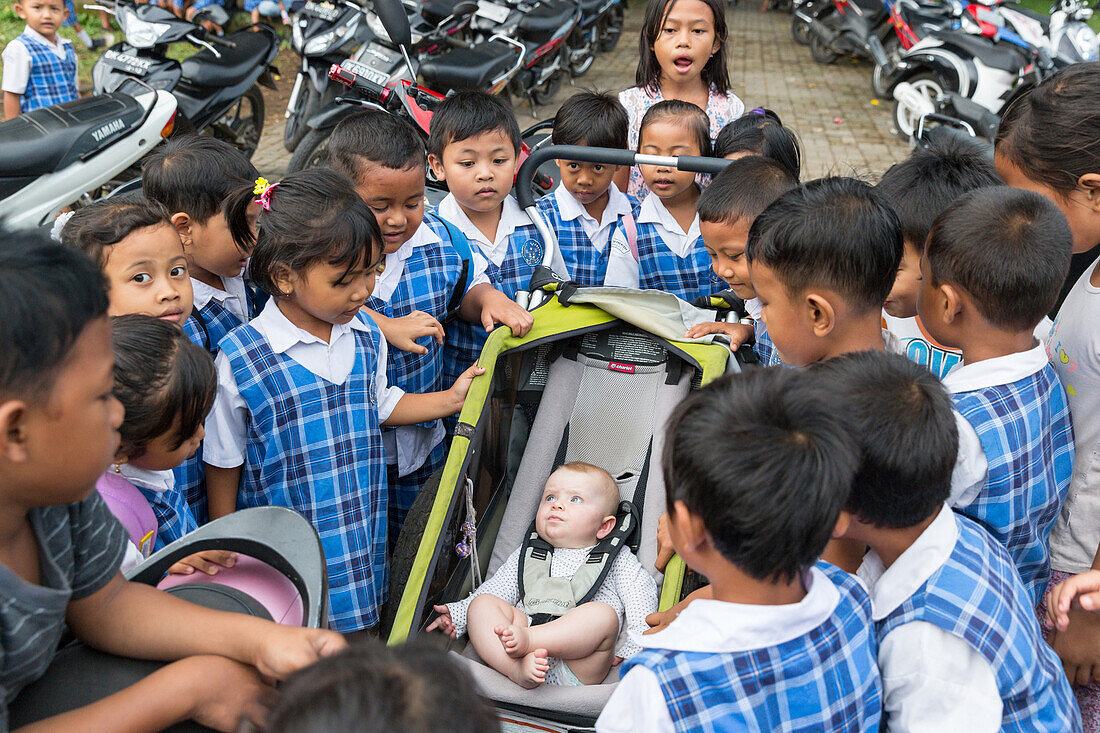 Many Balinese schoolchildren being curious about western baby, girl 5 months old, sitting in a stroller, kids gathering around, intercultural contact, meeting locals, family travel in Asia, parental leave, MR, Ubud, Bali, Indonesia