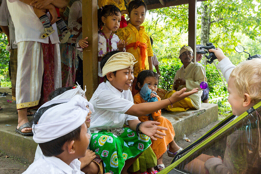 Balinese kids playing with German young boy, 3 years old, traditional clothes at temple ceremony, Balinese religion, stroller, intercultural contact, meeting local people, locals, family travel in Asia, parental leave, German, European, MR, Munduk, Bali, 