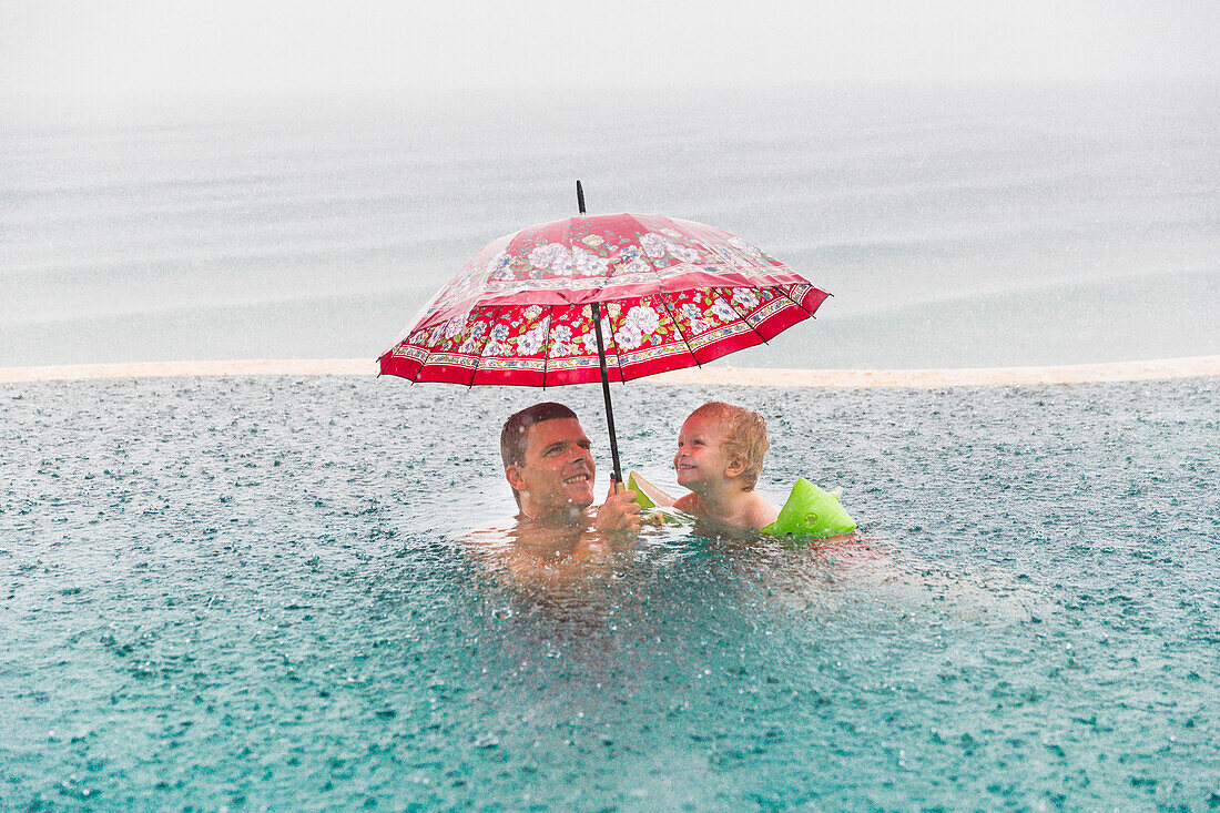 Father and son in a swimming pool, heavy rain, red umbrella, overflow pool, infinity pool, happiness, tropical rain, rainfall, sea, ocean, luxury, family travel in Asia, parental leave, German, European, MR, Amed, Bali, Indonesia