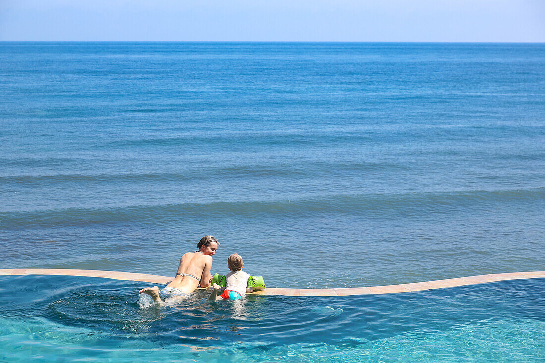 Mother and son in a swimming pool, overflow pool, infinity pool, looking to the sea, happiness, sea, blue sky, luxury, paradise, family travel in Asia, parental leave, German, European, MR, Amed, Bali, Indonesia