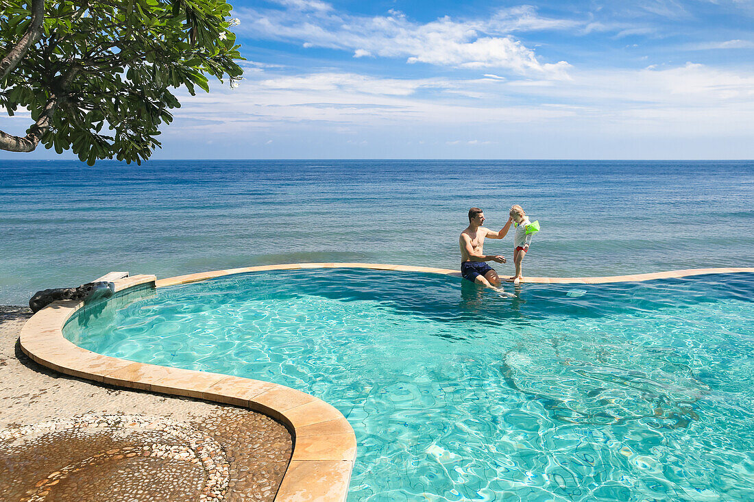 Father and son in a swimming pool, overflow pool, infinity pool, sitting on the rim, wall, tree, happiness, sea, tree, blue sky, luxury, paradise, family travel in Asia, parental leave, German, European, MR, Amed, Bali, Indonesia