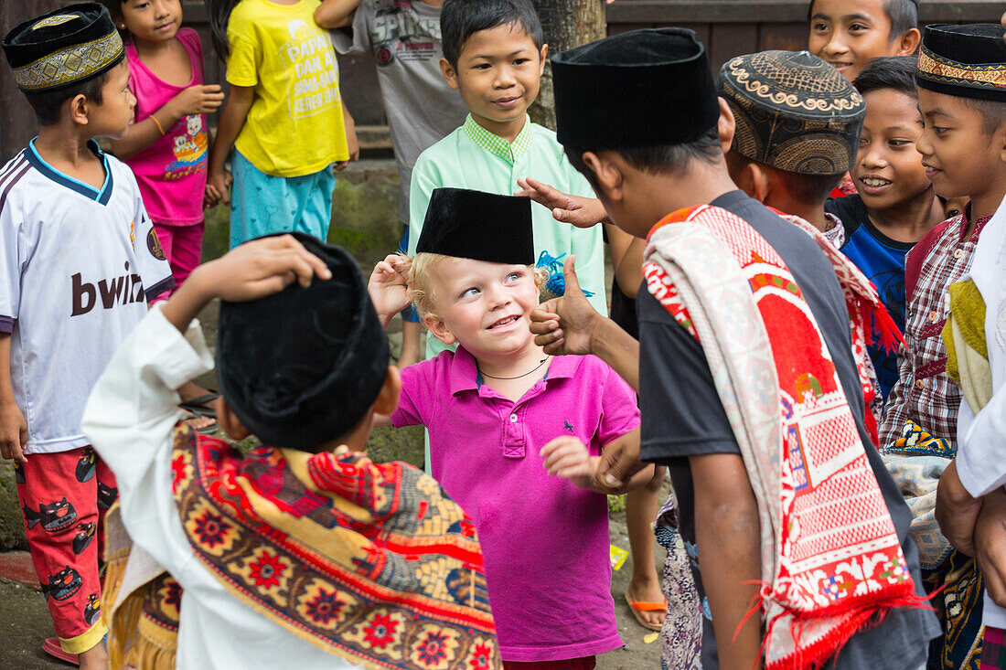 German little boy playing with Indonesian kids, children, boy 3 years old, trying their hat, fez, Islam, thumbs up, contact with local people, intercultural, family travel in Asia, parental leave, German, European, MR, Tetebatu, Lombok, Indonesia