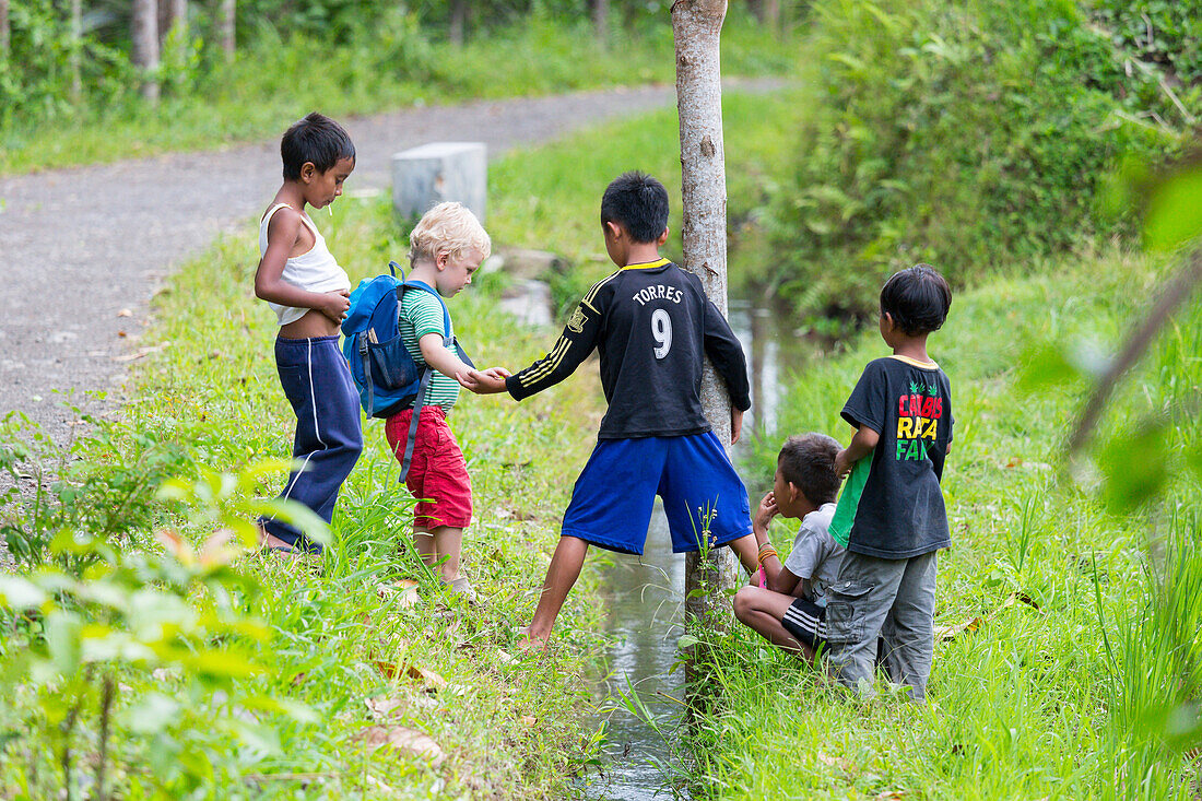 German little boy playing with Indonesian kids, crossing over a ditch, trench, giving a helping hand, soccer t-shirts,  children, countryside, village, boy 3 years old, contact with local people, intercultural, family travel in Asia, parental leave, Germa