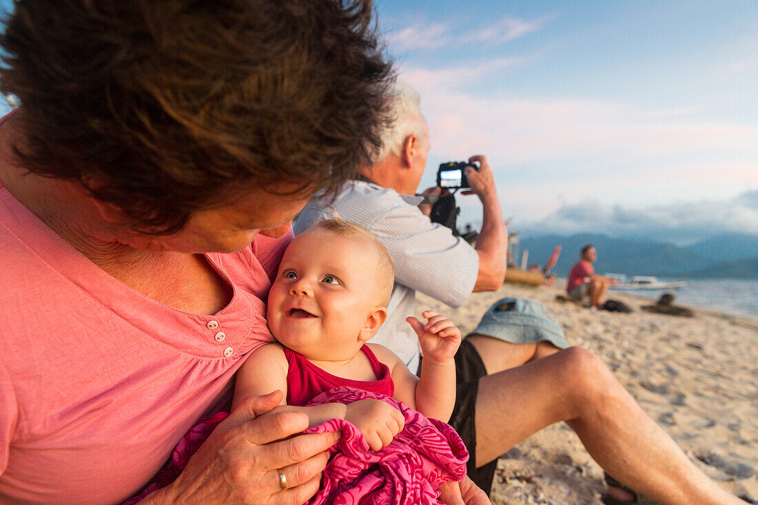 Grandmother with her granddaughter at the beach, sunset, laughing, smile, baby 5 months old, girl, grandfather taking pictures, tropical island, family travel in Asia, parental leave, German, European, MR, Gili Air, Gili Inseln, Lombok, Indonesia