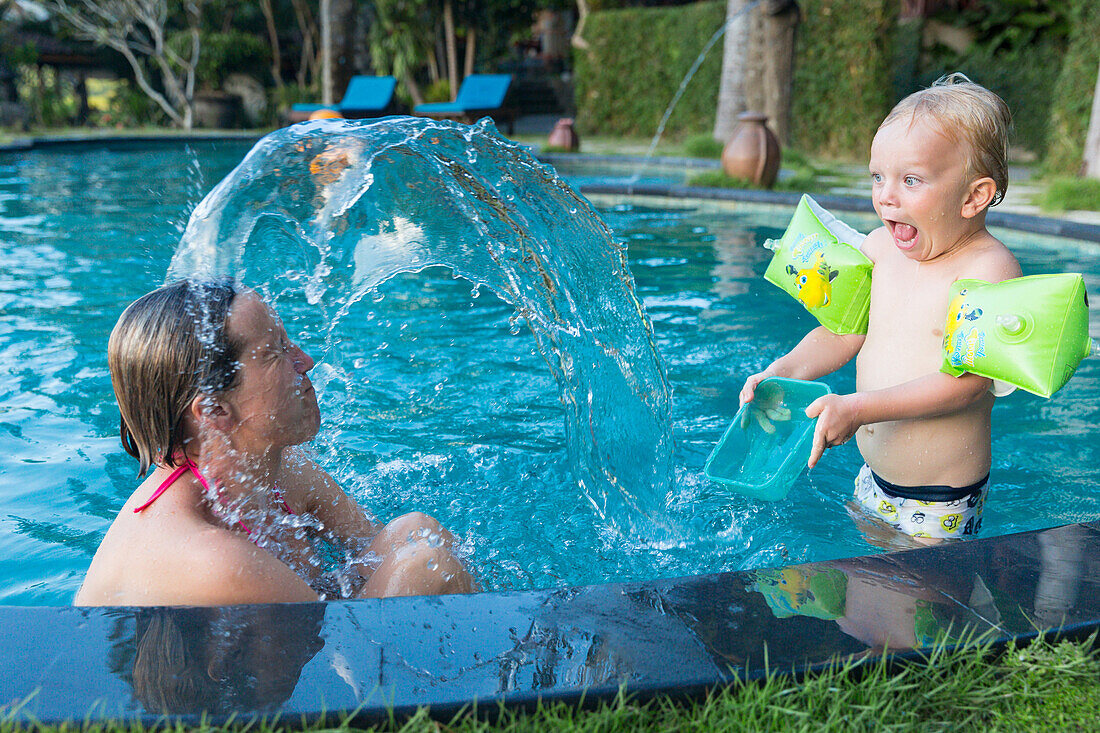 Mother and son playing in a swimming pool, son splashing water at his mum, surprise, swimmies, armbands, 3 years old, Balinese holiday resort, family travel in Asia, parental leave, German, European, MR, Sidemen, Bali, Indonesia