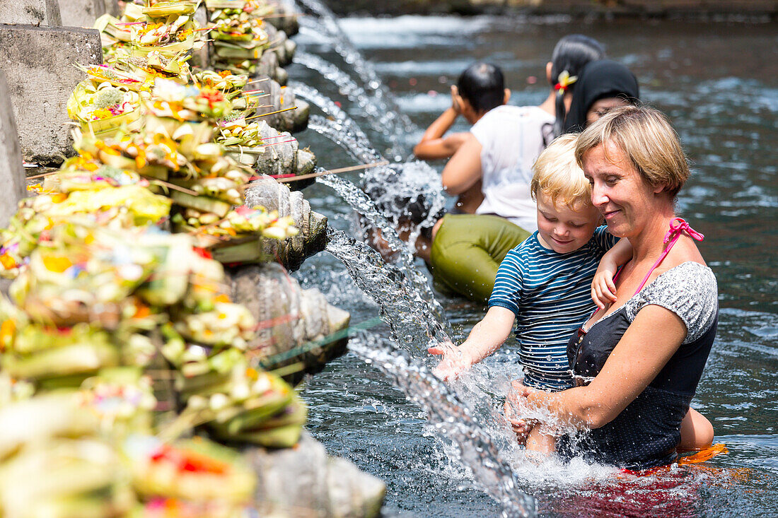 Holy springs Pura Tirta Empul, mother with her son on her arm taking a bath, pool, splashing water, water spilling pipes, tourists, temple, intercultural contact, locals, family travel in Asia, parental leave, German, European, MR, Ubud, Bali, Indonesia