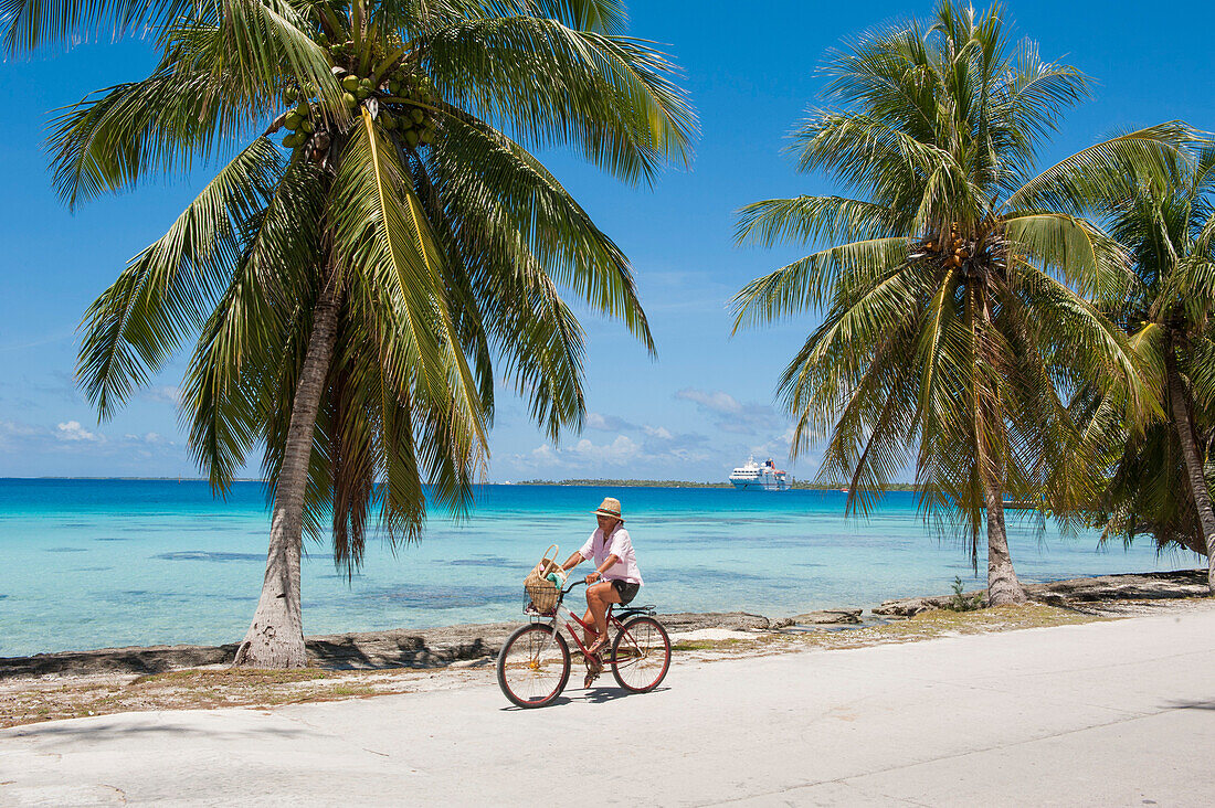 Mann fährt Fahrrad an einer Straße mit Palmen sowie Expeditions-Kreuzfahrtschiff MS Hanseatic (Hapag-Lloyd Kreuzfahrten) auf Reede im Hintergrund, Fakarava, Tuamotu-Inseln, Französisch Polynesien, Südpazifik