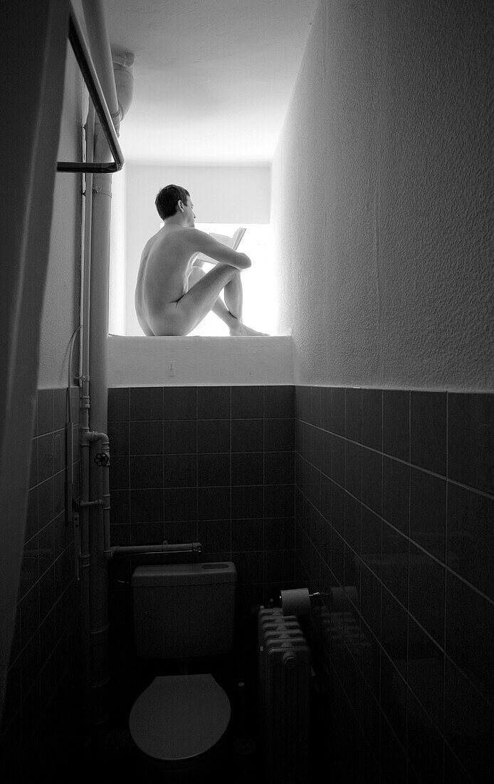 A naked man is sitting in front of an open window in his bathroom and reading a book (black and white photo), Berlin, Germany, Europe