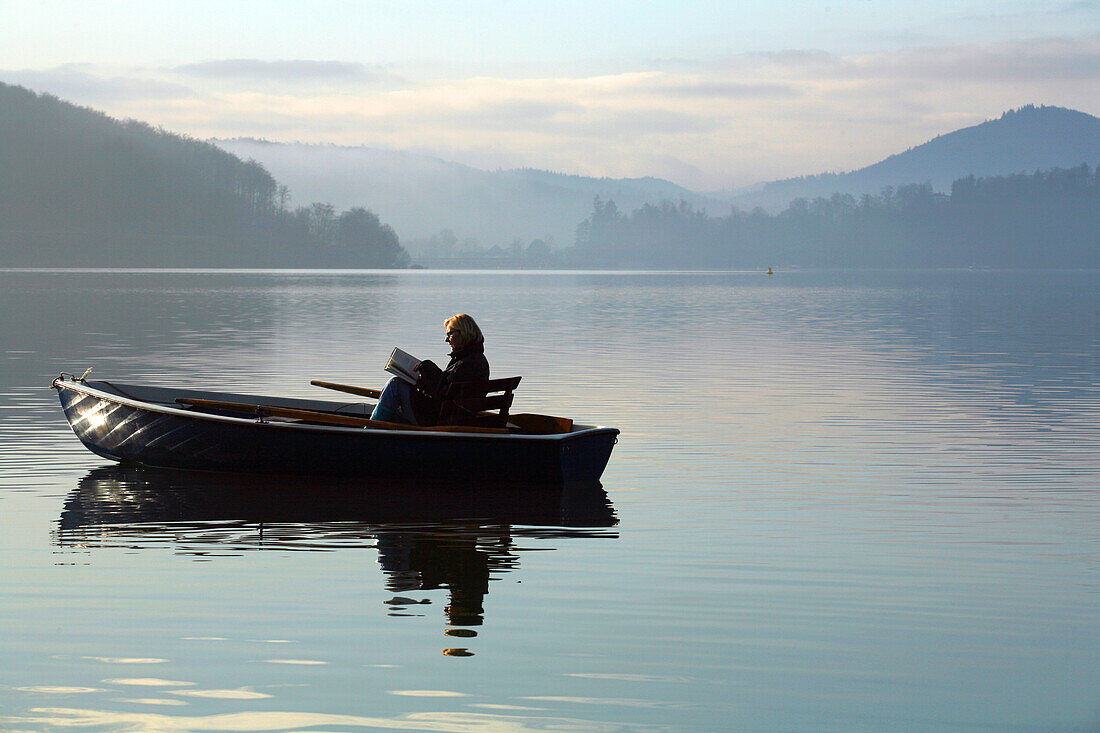 A woman sitting in a small dinghy fishing boat on Lake Edersee in Kellerwald-Edersee National Park reading a book in the morning light, Lake Edersee, Hesse, Germany, Europe