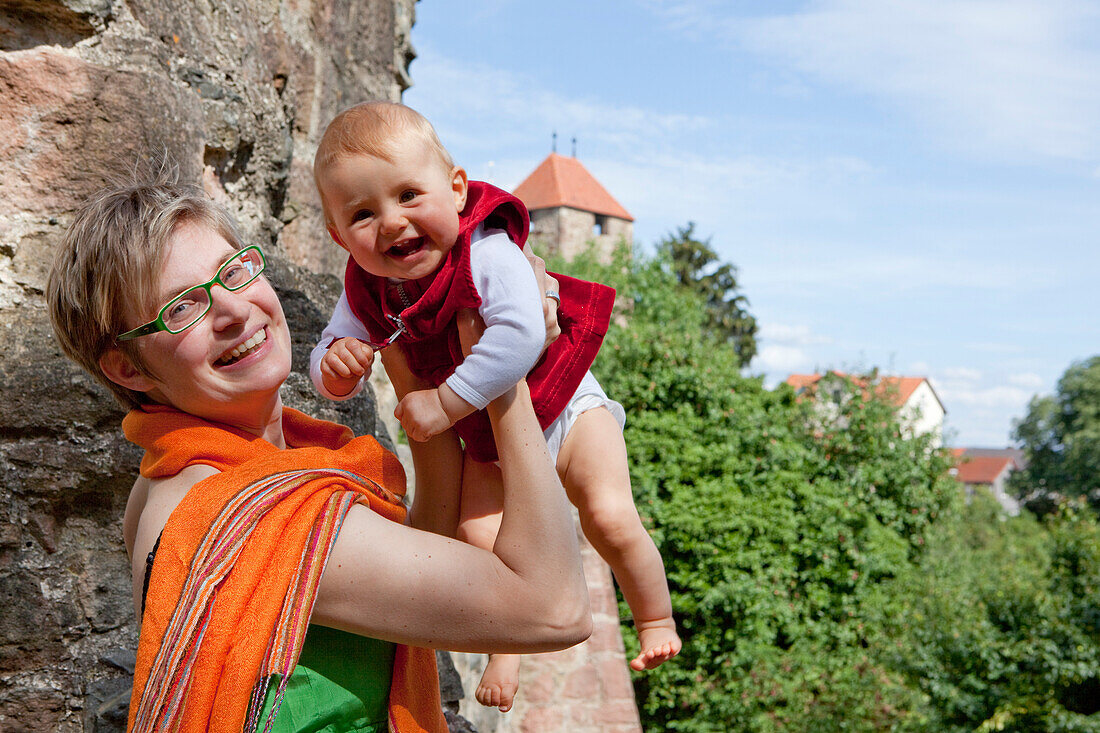 Junge lächelnde Frau mit grüner Brille hält ihr lachendes Baby an der Stadtmauer von Fritzlar hoch, Fritzlar, Nordhessen, Hessen, Deutschland, Europa