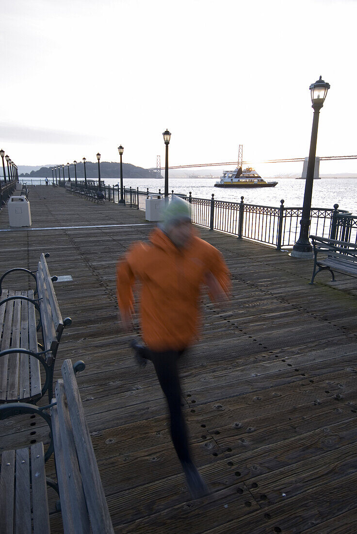 A man running on a dock in the harbour of San Francisco at sunrise with the Oakland Bay Bridge in the background. California, USA.  Lars Schneider / Aurora Photos 