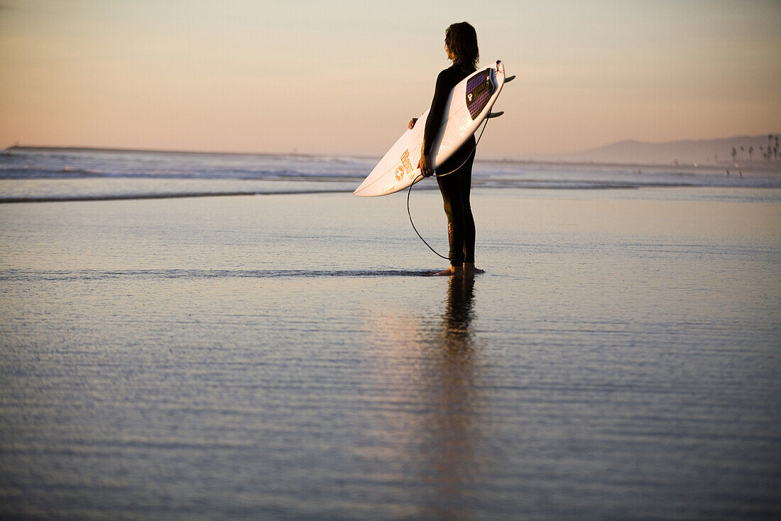 Surfer stands on the beach and looks out at the sunset in Oceanside, CA.